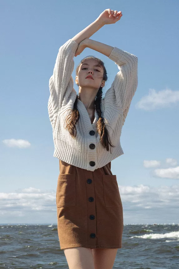 A woman wearing a sweater and skirt stands on the beach, with waves gently lapping at the shore in the background