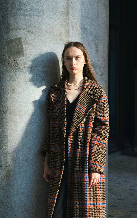 A woman in a coat leans against a wall, looking contemplative in a casual urban setting