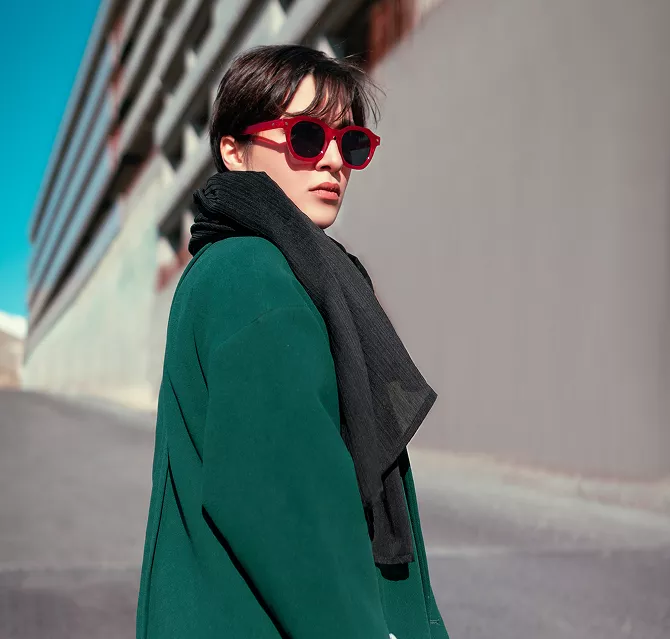 A woman in sunglasses and a green coat stands confidently, showcasing her stylish outfit against a neutral background
